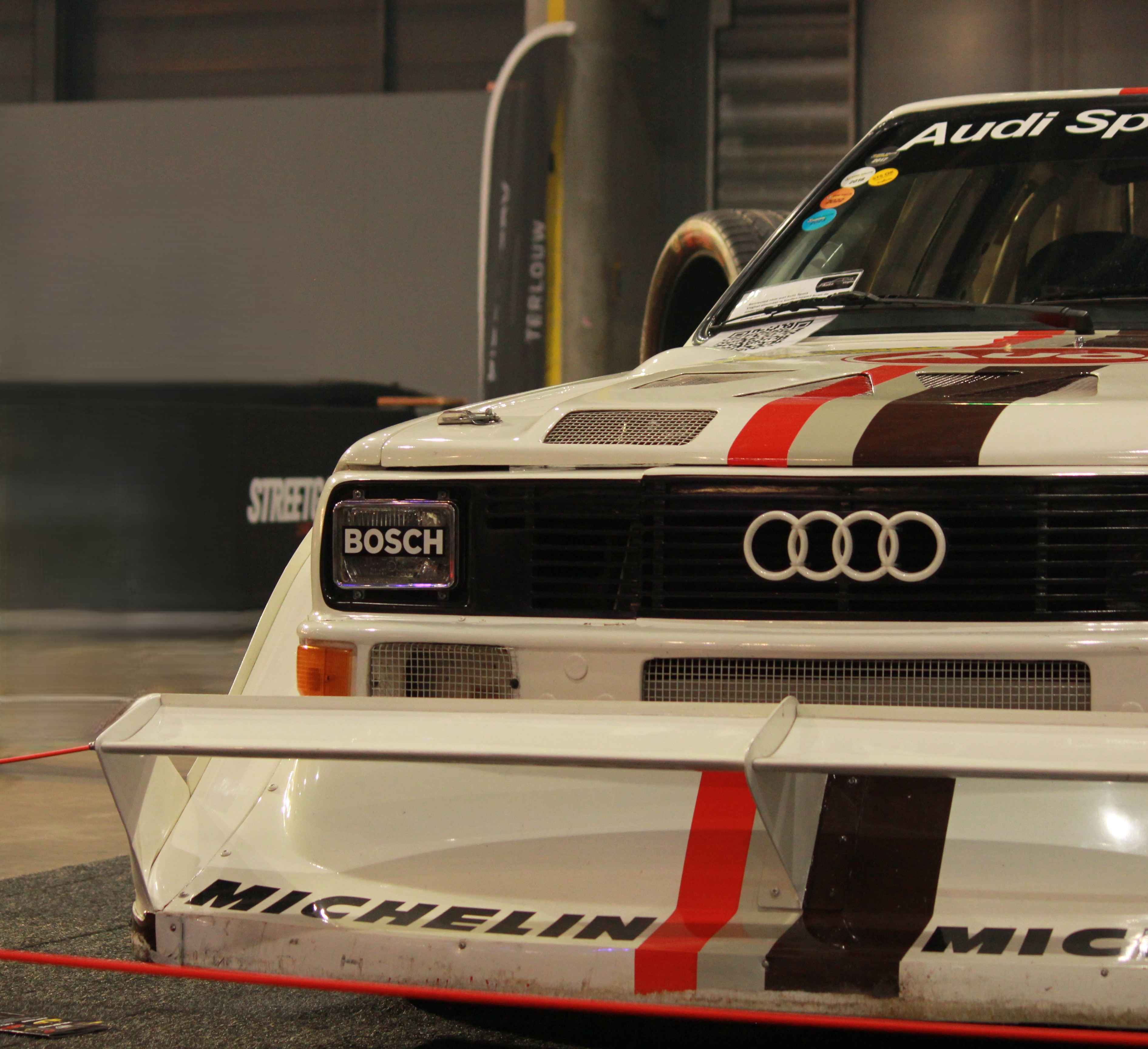 Front view of a classic Audi Quattro rally car with Bosch and Michelin branding, displayed indoors at a car show.